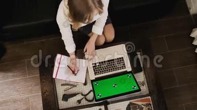 一个金发女孩带着一台笔记本电脑，她在笔记本上工作并做笔记。 木制内部。 绿色笔记本电脑